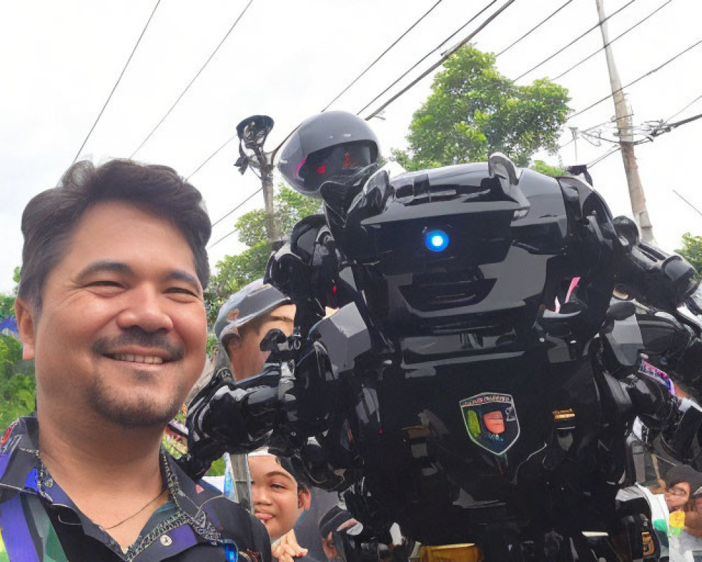 Smiling man with patterned shirt next to life-size black robot at outdoor event