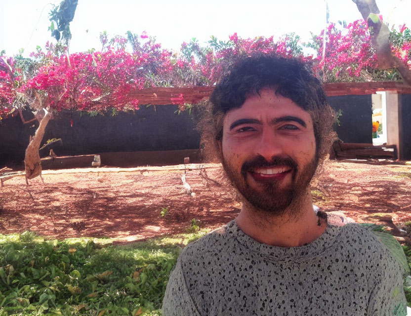 Bearded man smiling outdoors with pink flowers and wooden trellis