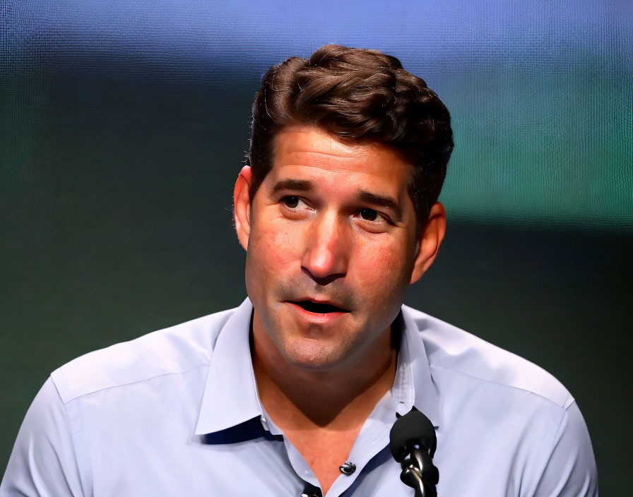 Dark-Haired Person in Light Blue Shirt Speaking into Microphone on Green Bokeh Background