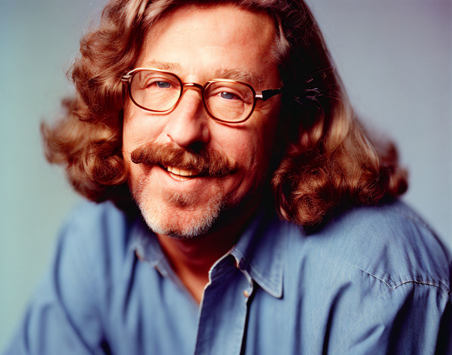 Smiling man with curly hair and glasses in denim shirt on blue background