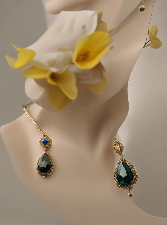 Mannequin bust showcasing gold earrings with green and blue gemstones and a white flower accent.