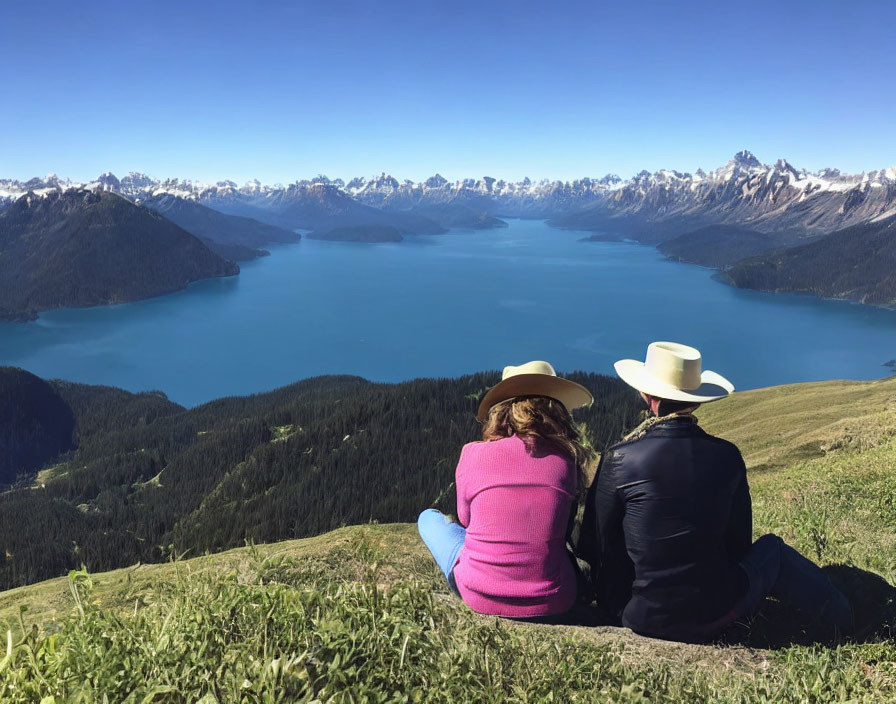 Cowboy-hat-wearing duo on grassy hill by blue lake and mountains