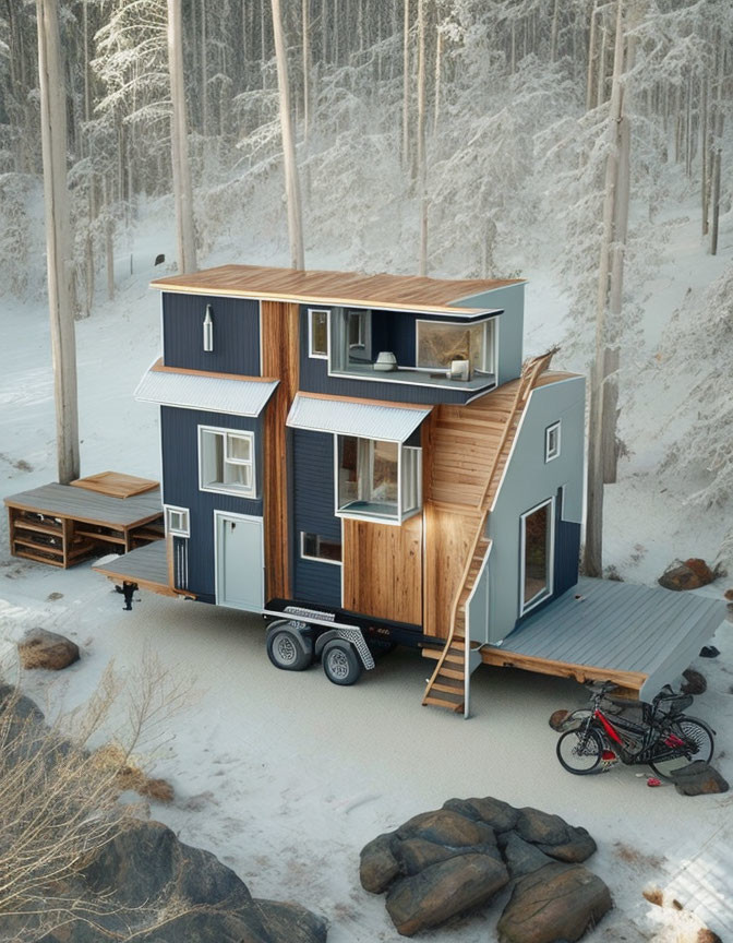 Two-story tiny house on wheels in snow-covered forest with wood and dark blue siding, wooden deck,