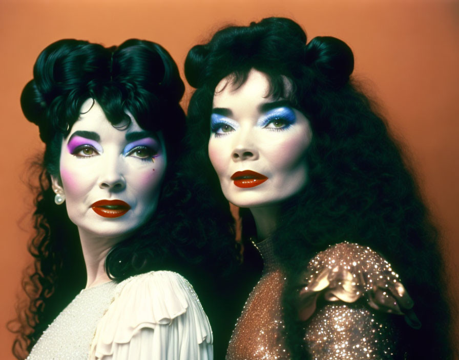 Two women in dramatic makeup and vintage hairstyles in glittery outfits on orange backdrop