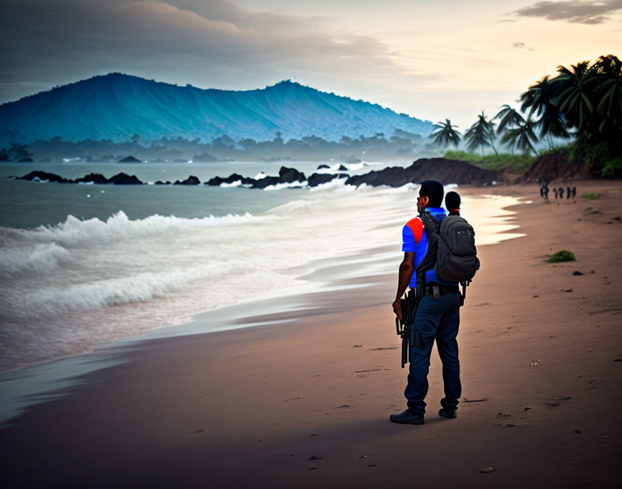 Person in Blue Uniform on Sandy Beach with Mountains and Palm Trees