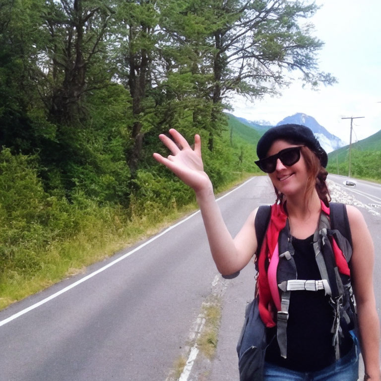 Smiling woman with backpack, hat, and sunglasses waving on roadside