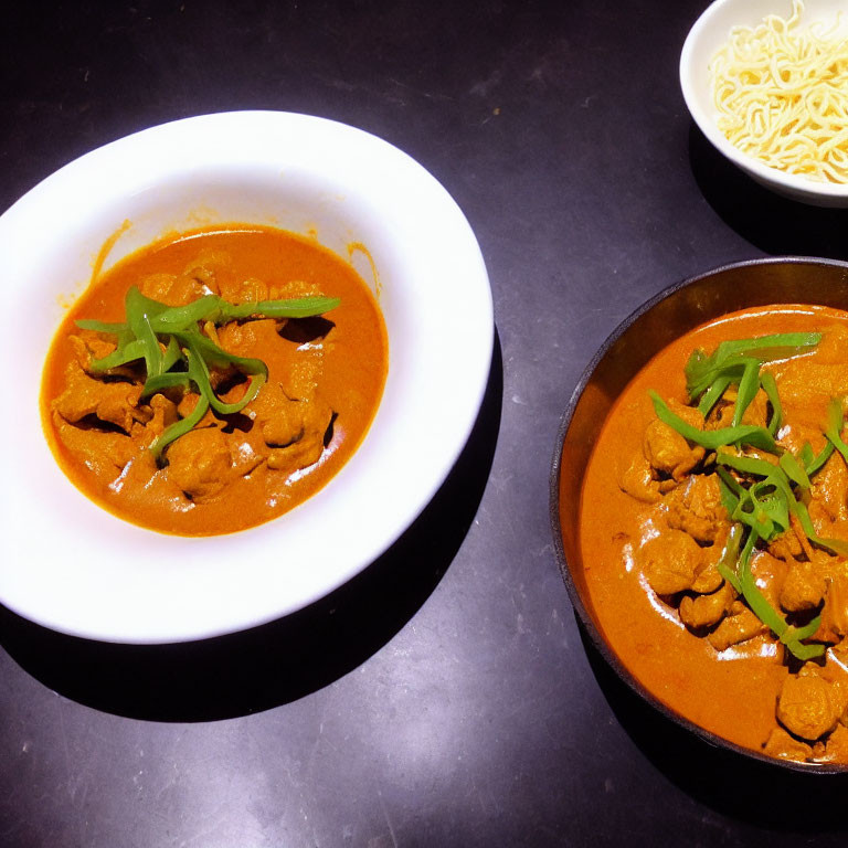 Bowls of Curry with Meat and Green Garnish Next to Noodles on Dark Table