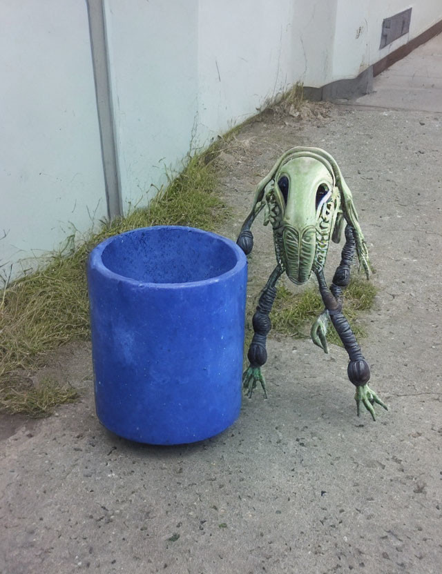 Green alien-like digital creature with mechanical limbs beside blue trash can