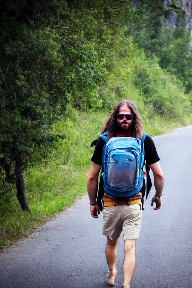 Bearded person with sunglasses and backpack walking by forest on road