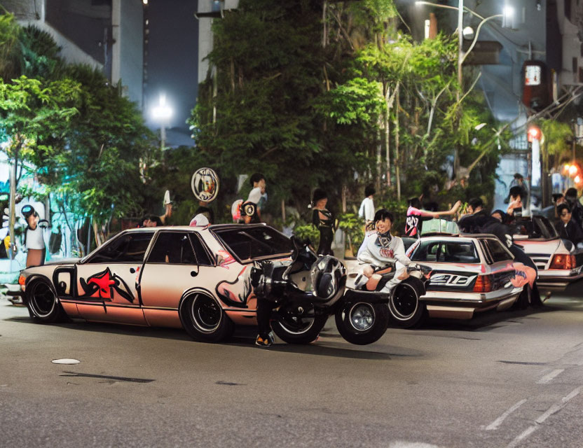 Group of people admiring modified pink and black cars on city street at night