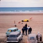 Three people under striped umbrella on wooden walkway by beach with caravan and boat.