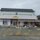 Abandoned two-story house with peeling paint amid debris under cloudy sky