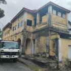 Abandoned two-story house with overgrown plants, rusted car, and debris in foggy landscape