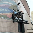 Tiled facade with black cutouts, obscured by trees and scaffold