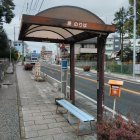 City street bus stop with metal shelter, cars passing, and residential buildings.