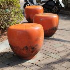 Orange and white apple-shaped chairs on beige surface with fallen petals, by flower wall under sunny sky