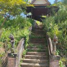 Triangular facade rustic house in lush green landscape with stone steps, balcony, and natural boulders