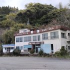 Weathered two-story building with peeling paint, boarded windows, balcony, and overgrown vegetation