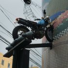 Figure in leather jacket balancing floating motorcycle surrounded by fluttering leaves, child and hovering figure in orange observe