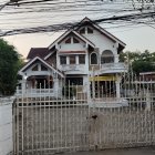 Two-story house with gabled roof, balconies, white railings, surrounded by greenery.