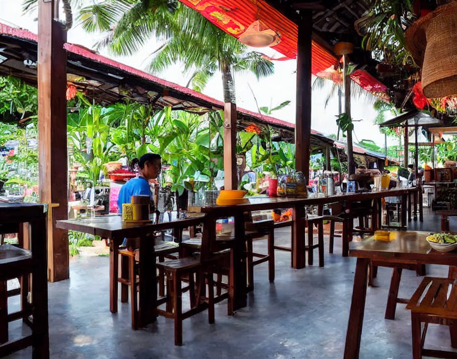 Person sitting at wooden bar in tropical outdoor restaurant with palm trees and red lanterns.