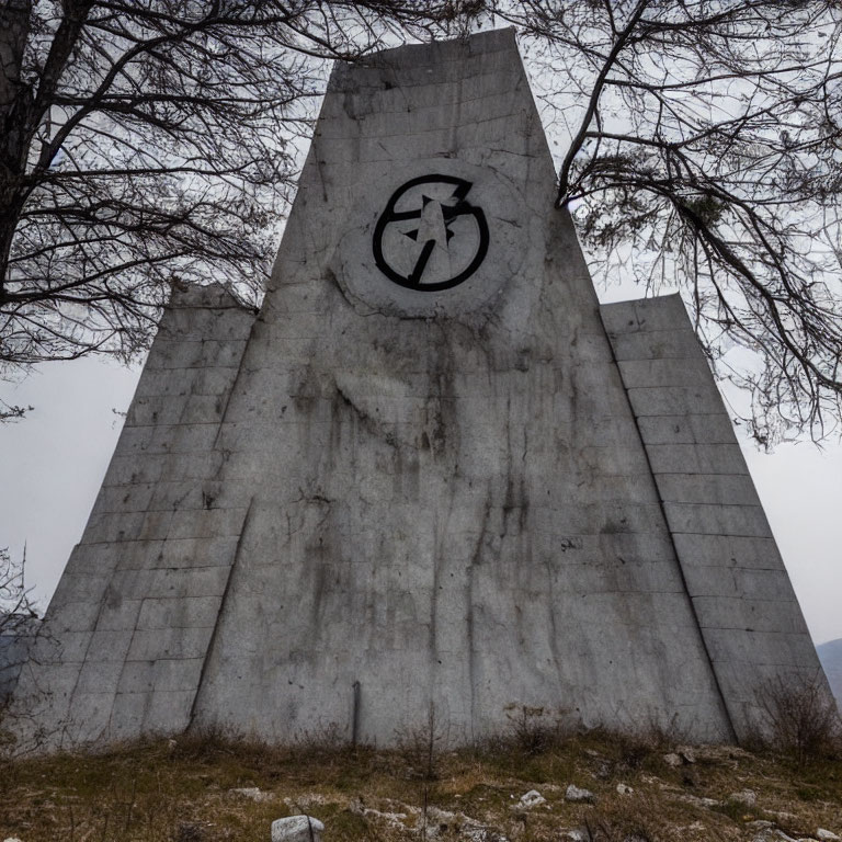 Weathered concrete monument with cyclist symbol and bare trees under cloudy sky