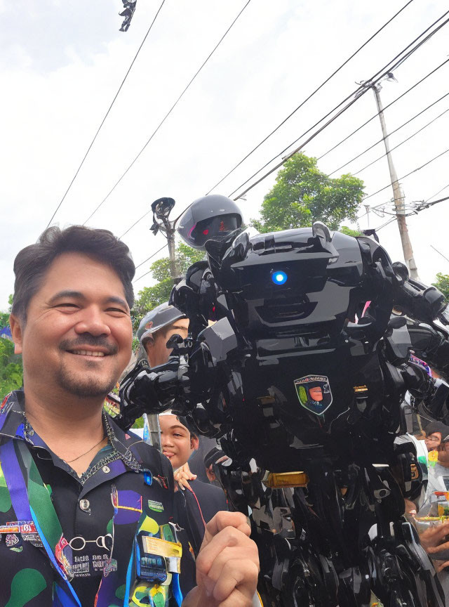 Smiling man with patterned shirt next to life-size black robot at outdoor event