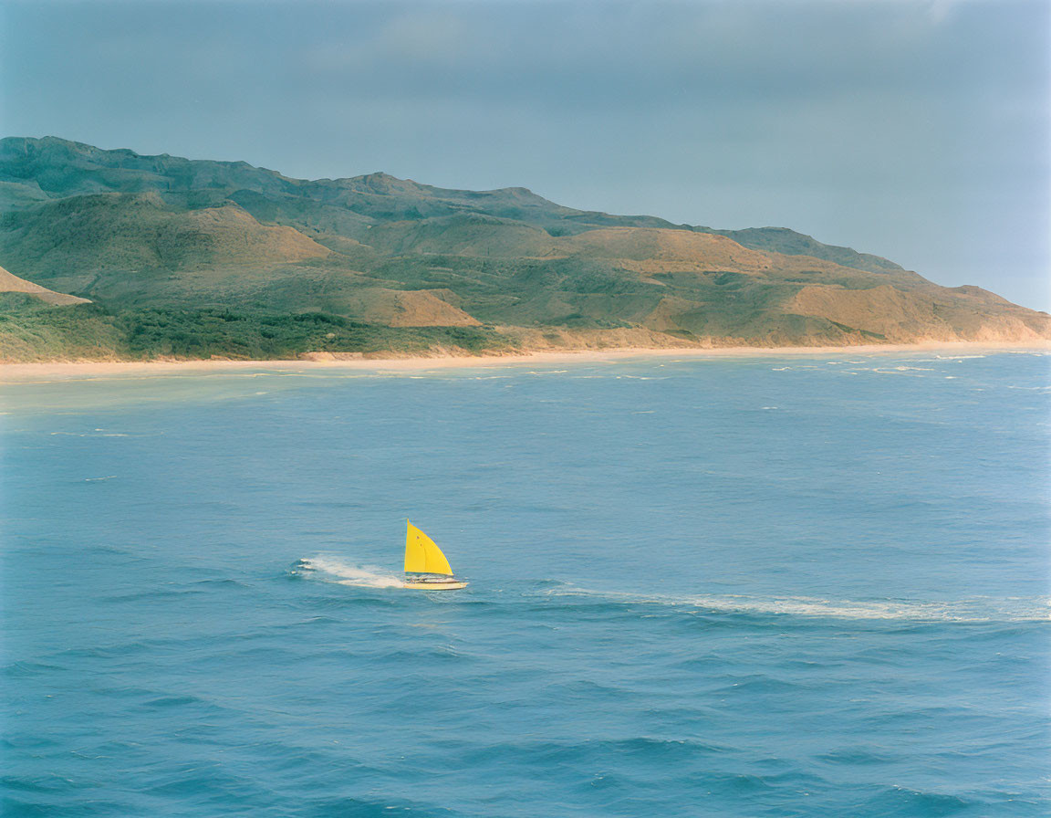 Yellow sail sailboat cruising near mountainous coastline on clear blue sea