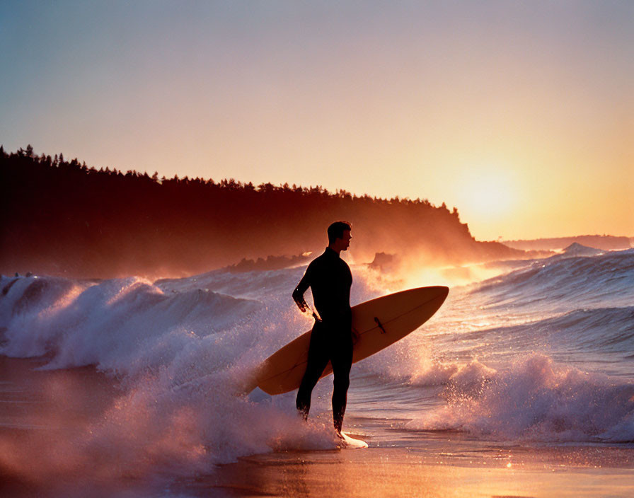 Surfer with board on beach at sunset, waves crashing, trees silhouetted.