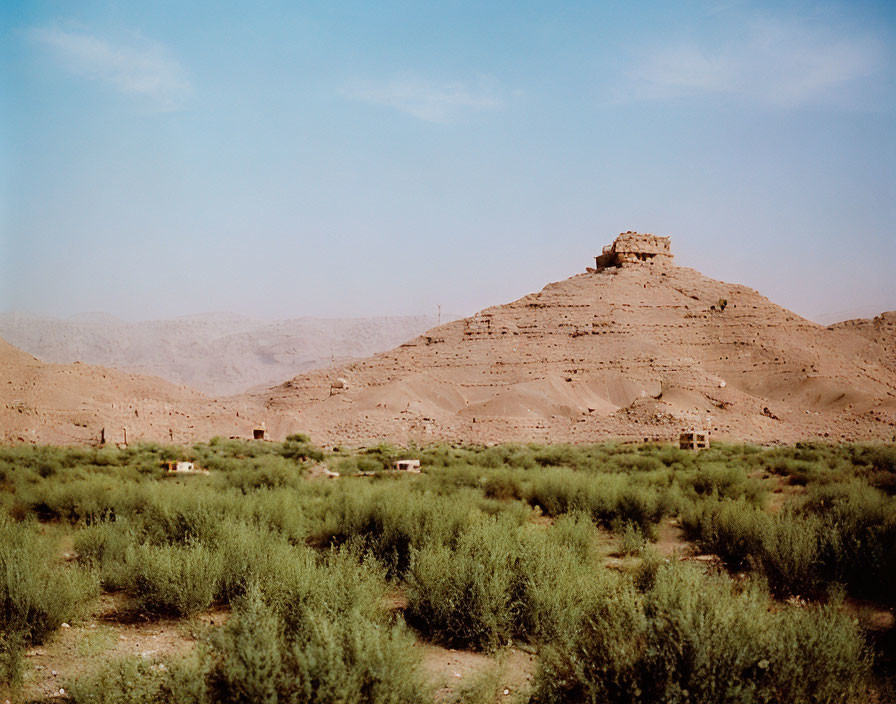 Sunlit arid landscape with small green shrubbery and ancient hilltop ruin under clear blue sky