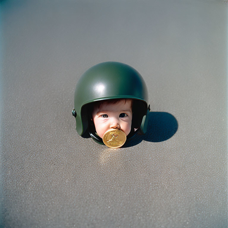 Baby wearing green helmet holds gold coin in mouth, face visible