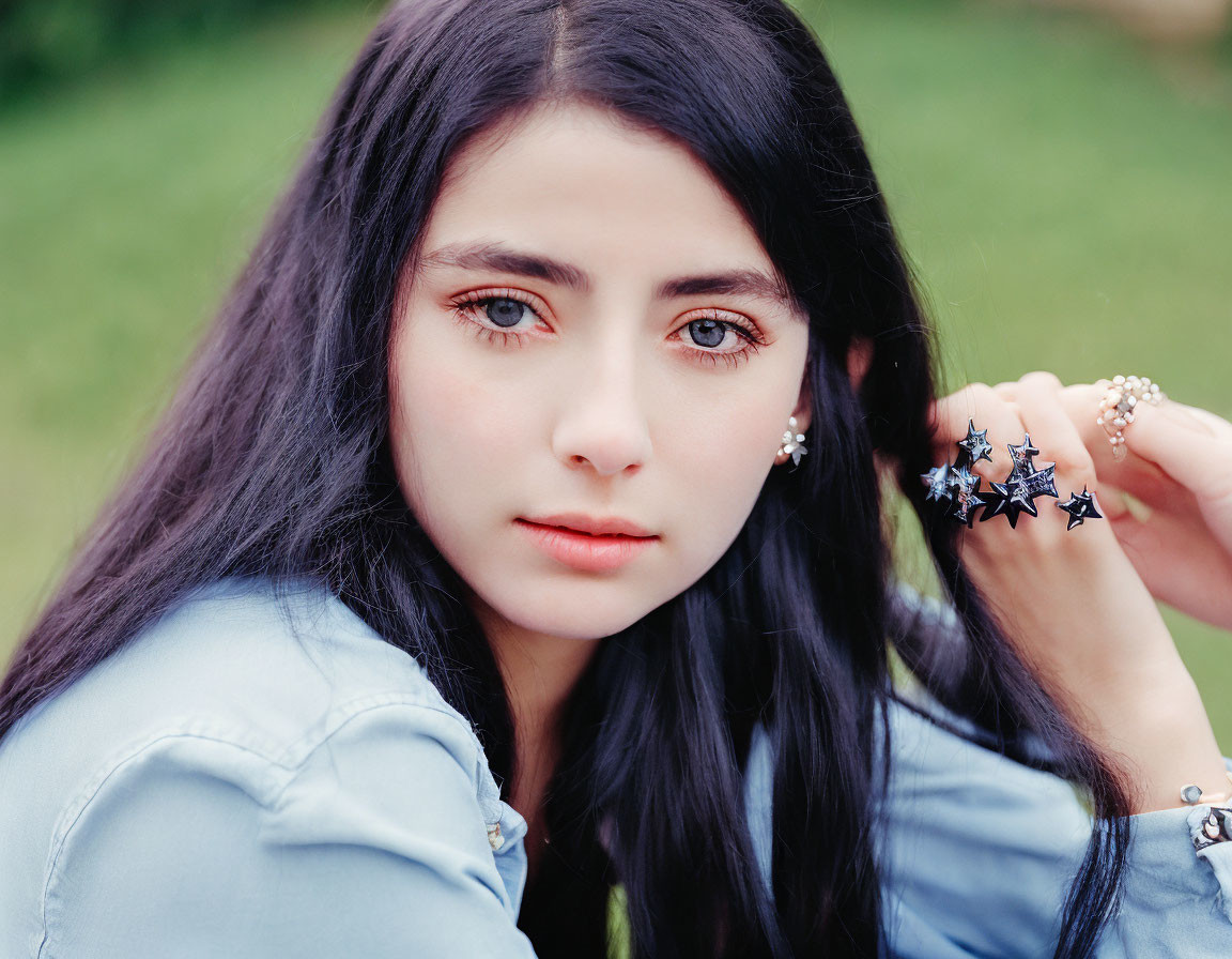 Young woman with long dark hair and blue eyes in denim shirt and star earrings poses outdoors.