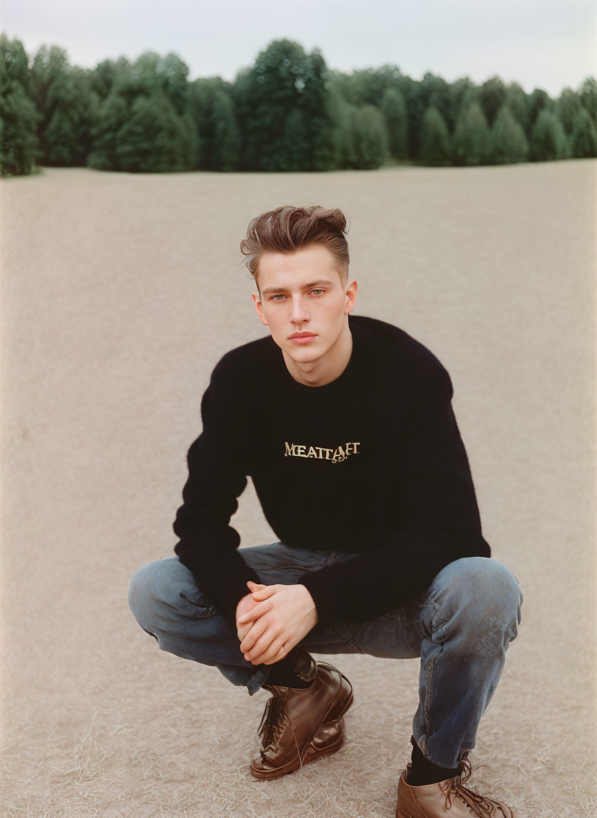 Young person in blue jeans and black sweater squatting on dry grass field with trees in background