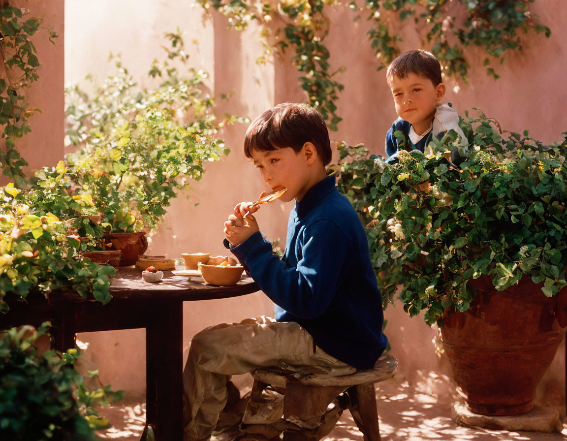 Two boys in a sunny courtyard, one eating at a small table under potted plants while the other