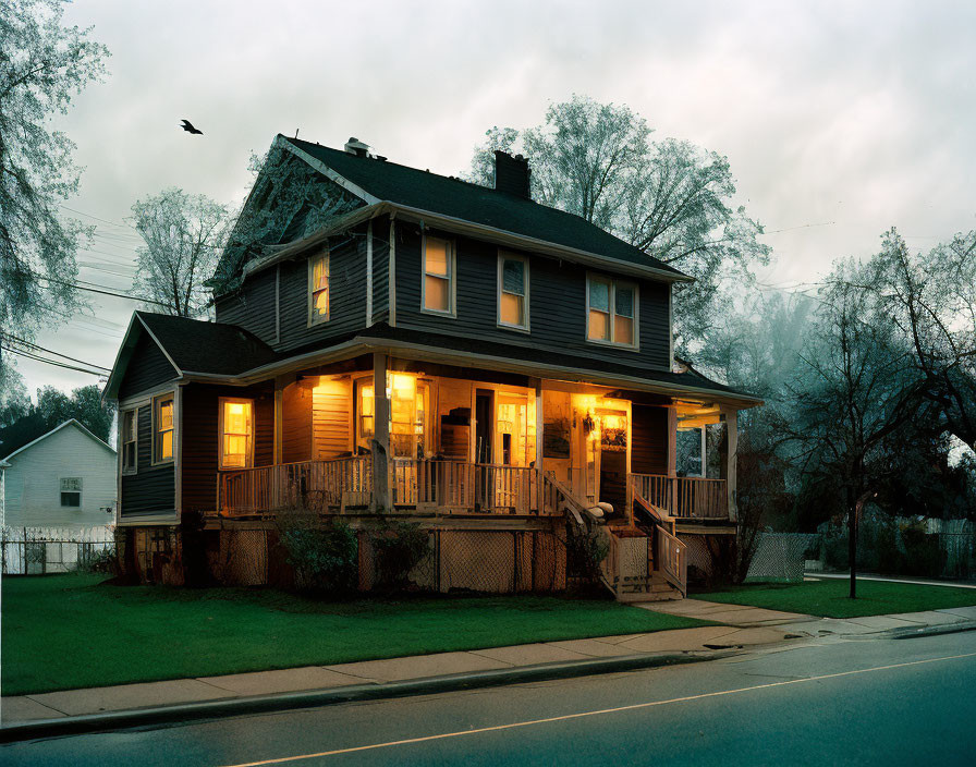 Twilight scene with illuminated two-story house, front porch, leafless trees, and bird in cloudy