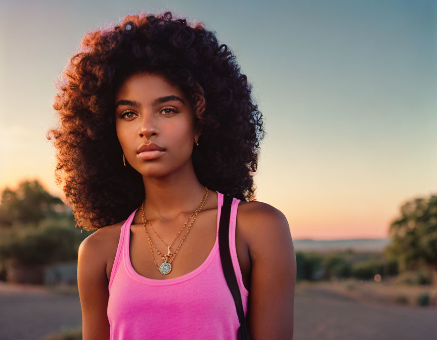 Young woman with voluminous curly hair in pink tank top outdoors at golden hour