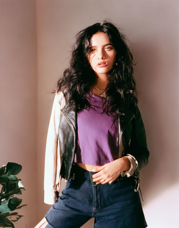 Casually posed woman in purple top, leather jacket, and blue jeans against neutral background with plant