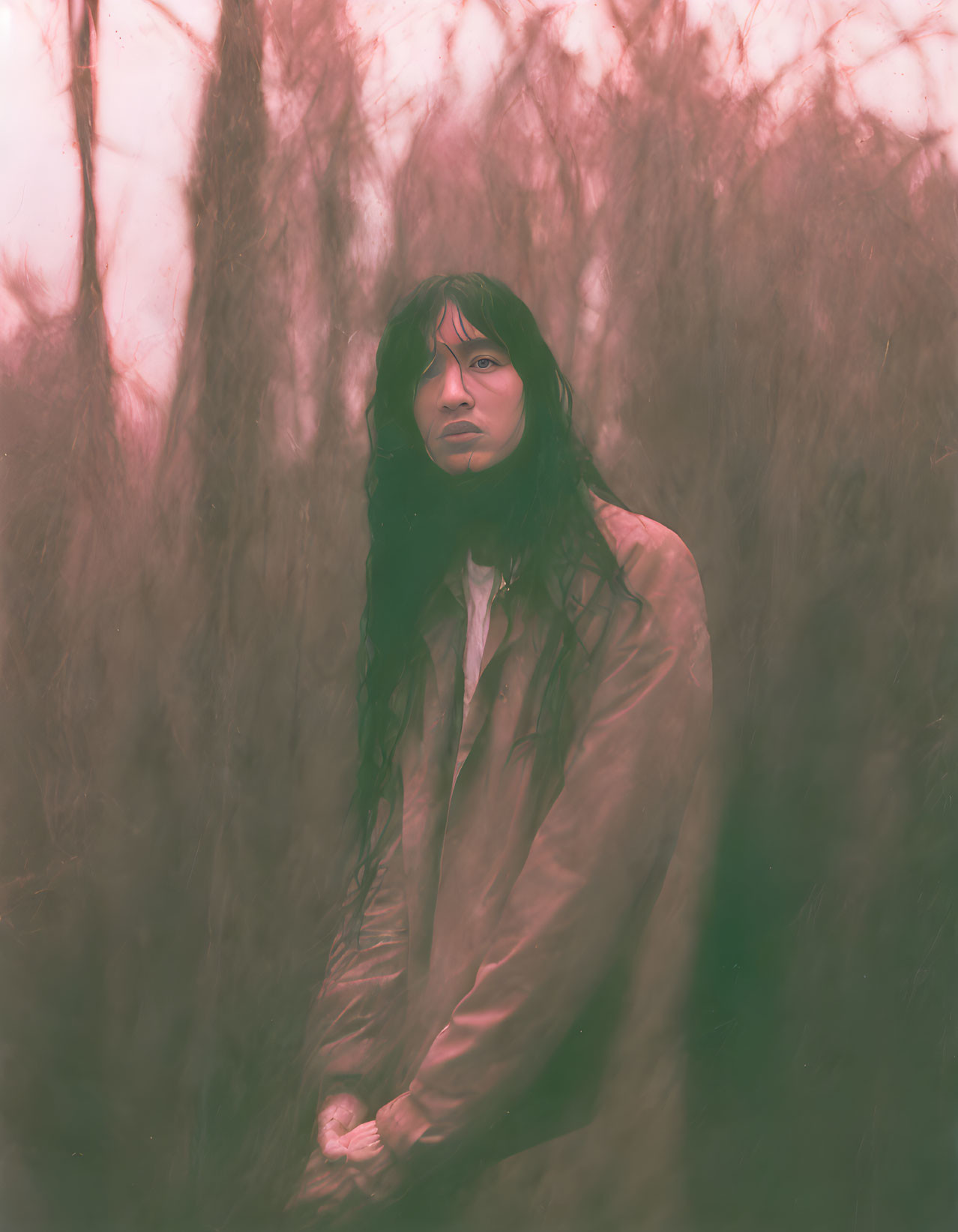 Long-haired person in misty, willow-draped setting with beige jacket exudes moody