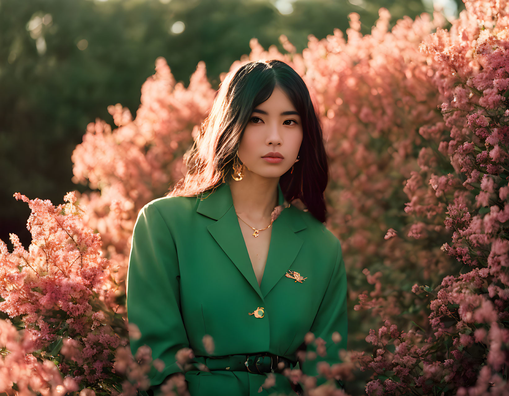 Dark-haired woman in green blazer surrounded by pink blossoms under warm sunlight