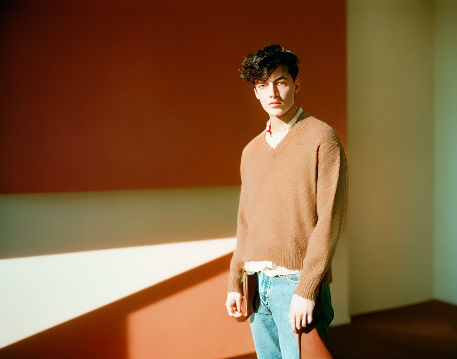 Young man in brown sweater and jeans standing in room with geometric shadows on red wall