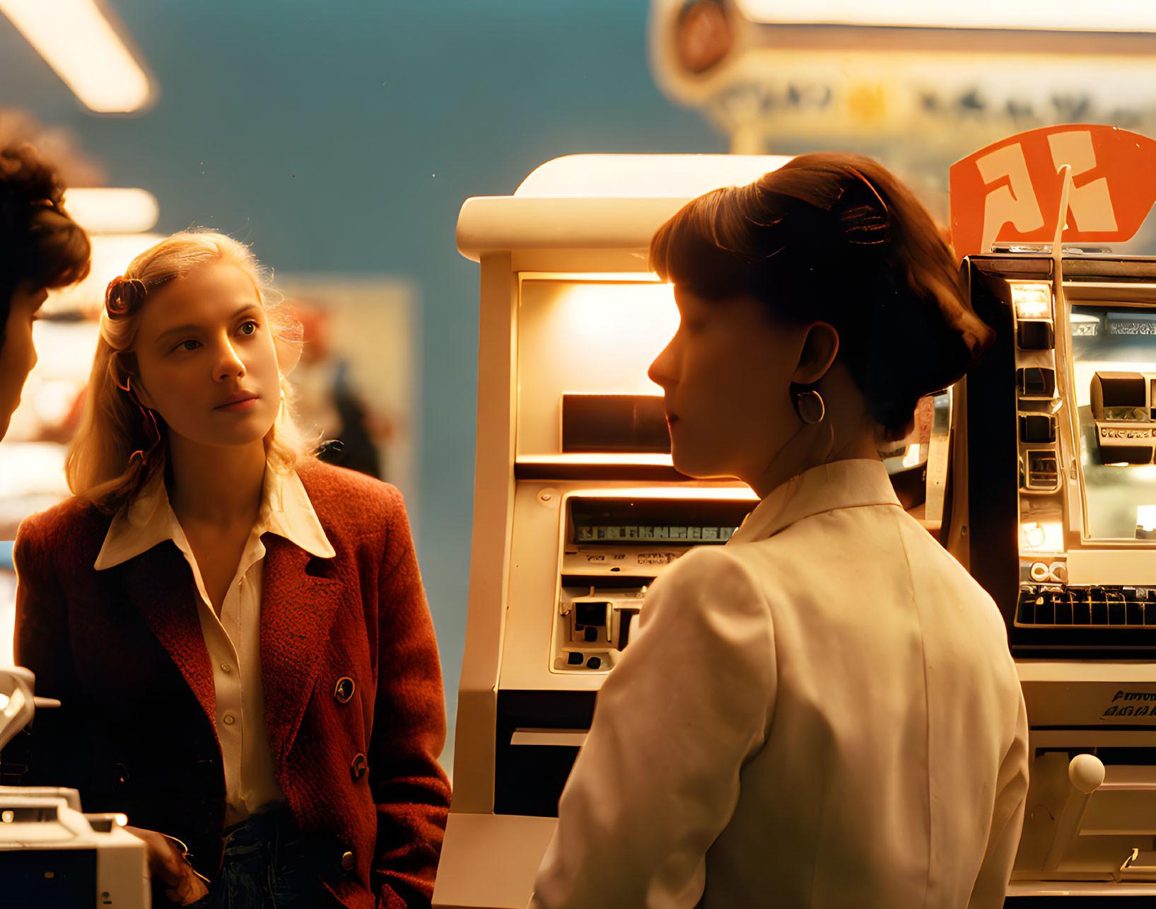 Young woman in red jacket listens to female cashier at retro checkout counter.