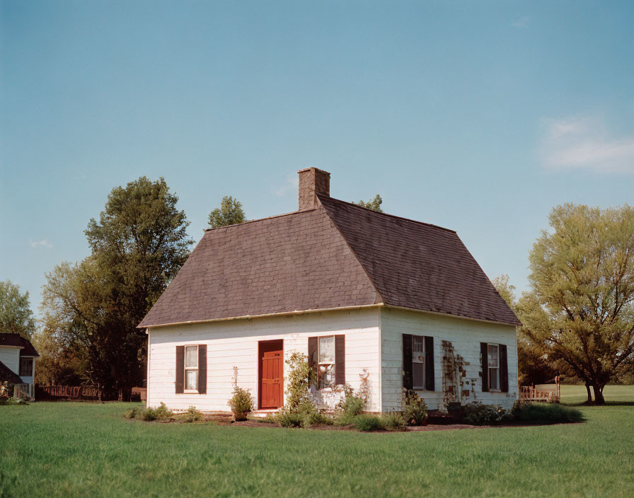 Vintage single-story house with shingled roof, red door, surrounded by greenery under blue sky