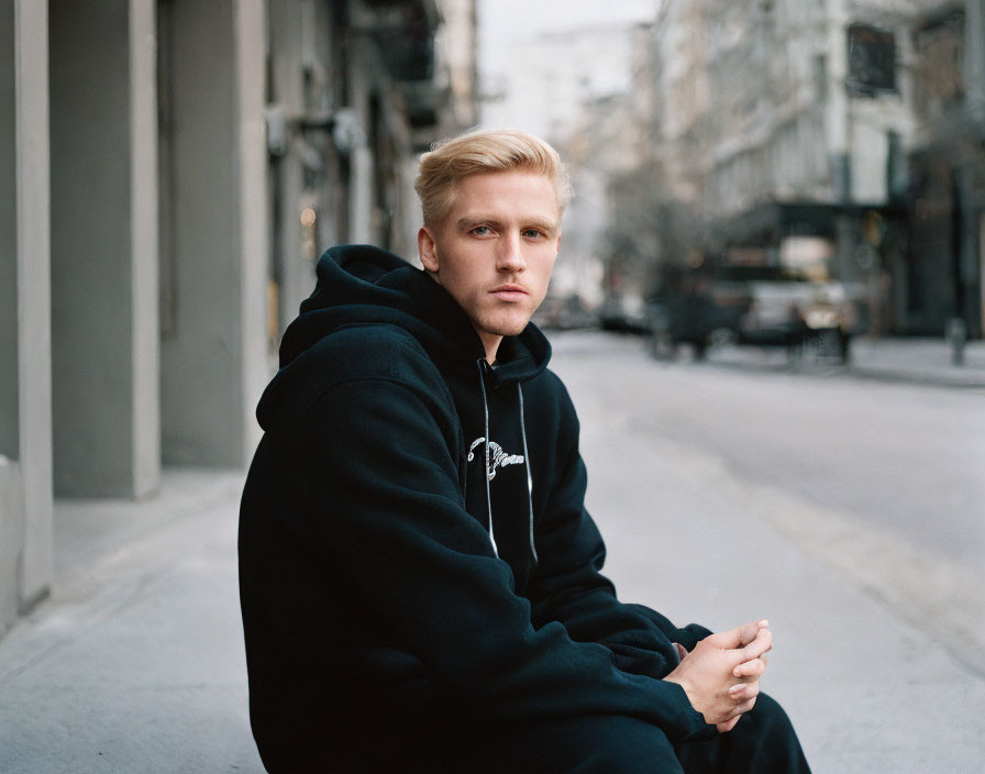 Blond-Haired Young Man in Black Hoodie on Curb with City Street Background