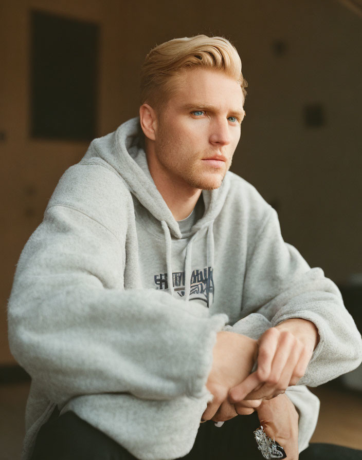 Blonde man in grey hoodie sitting with clasped hands, looking pensive