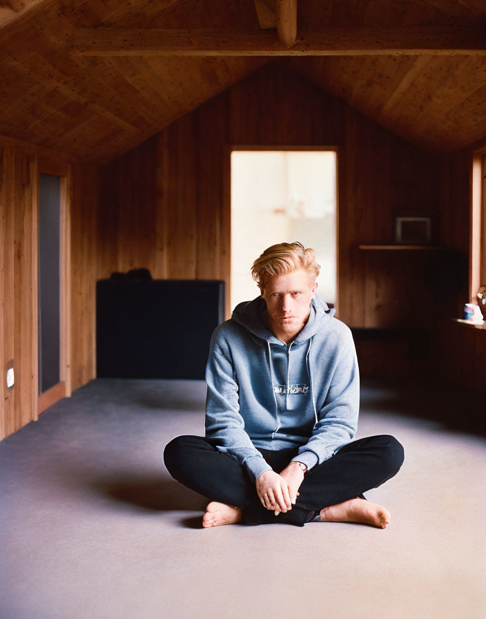 Person in blue hoodie sitting in attic room with wooden walls, looking pensive