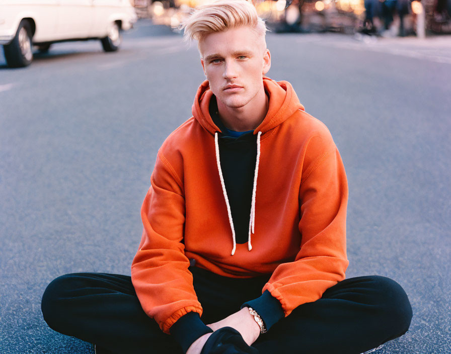 Blond-Haired Young Man in Orange Hoodie and Black Pants Sitting Cross-Legged