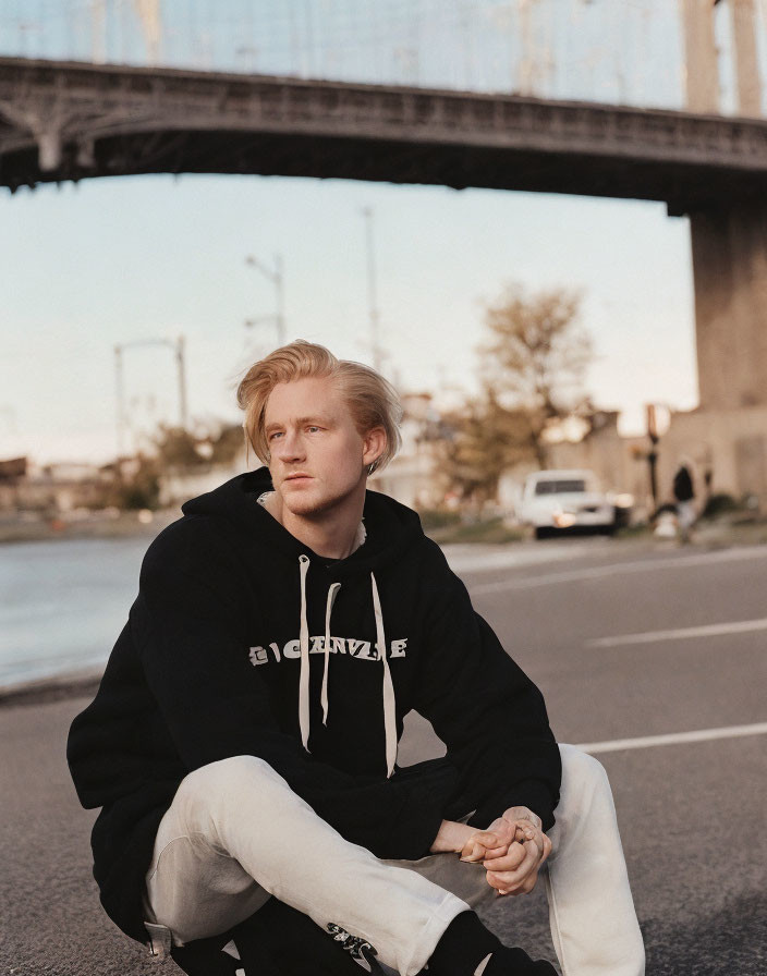 Blond person in black hoodie and jeans squatting on road with bridge at dusk