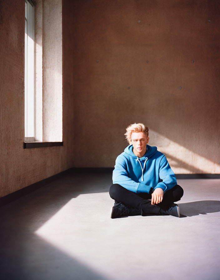 Blond Person in Blue Hoodie Sitting in Room with Large Window