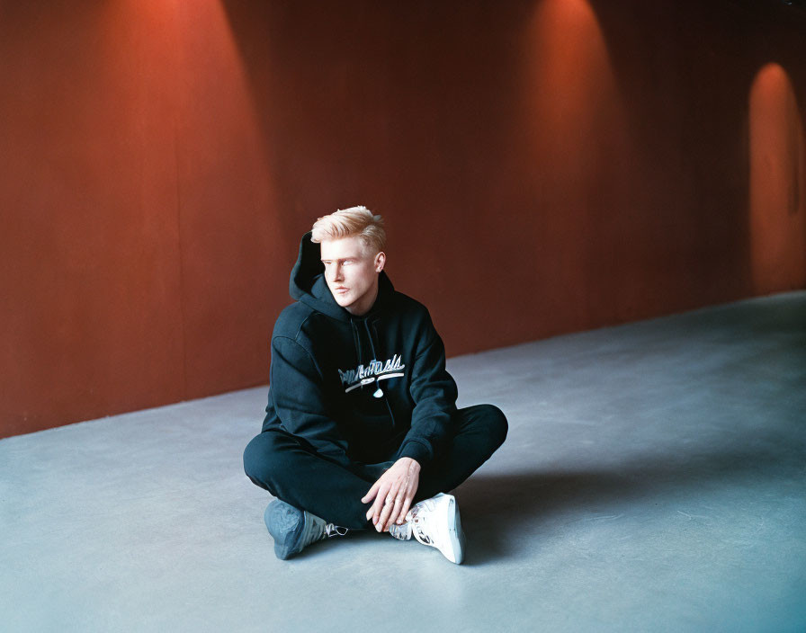 Blond person in black hoodie and white shoes against terracotta background