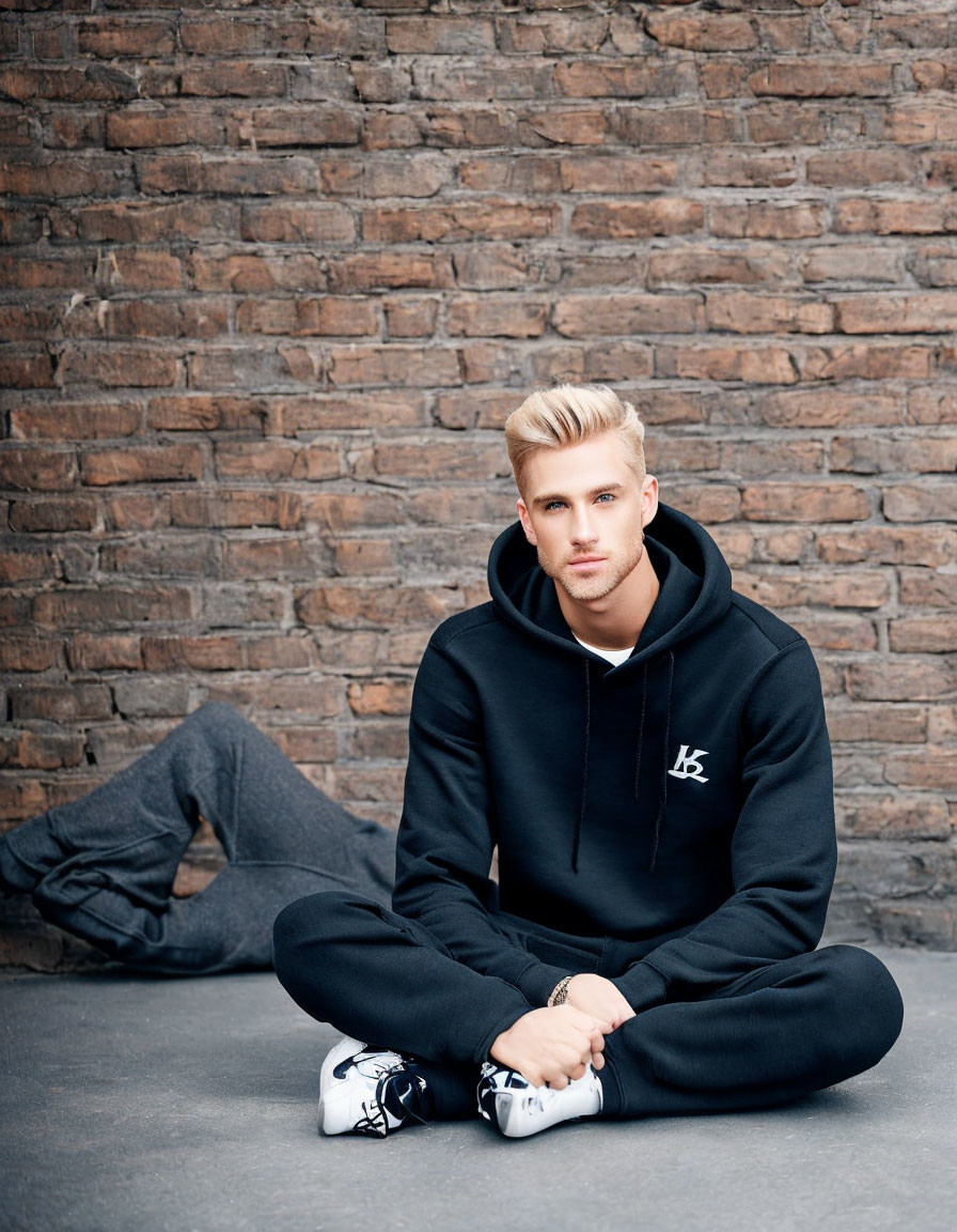 Blond man in black hoodie and sweatpants sitting cross-legged by brick wall